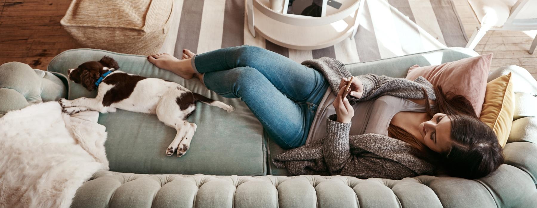 woman lays on a couch and texts next to her dog